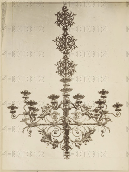 Chandelier decorated with leaf motifs, Luster en fer forgé et repoussé, photograph, page, name: 'Witz & cie Phot. Rouen', inscription: 'Composé et exécuté par F. Marrou, pour M. Depeaux à Rouen.', photographer: Witz et Cie., c. 1882 - in or before 1884, paper, albumen print, height 278 mm x width 215 mm, ornament derived from plant forms, chandelier, candelabrum, The image shows the upper portion of an ornate metal chandelier against a plain light background, featuring a central vertical rod with symmetrical scrollwork and decorative floral elements, multiple layered ornamental rosettes and leaf-like motifs, curled metal arms extending outward that support cup-shaped flower forms, and a repeating circular lattice of intricately twisted metalwork running horizontally across the piece.