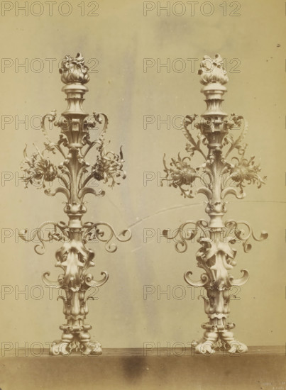 Two candlesticks decorated with leaf motifs and a flame, photograph, page, name: 'Witz & cie Phot. Rouen', photographer: Witz et Cie., c. 1874 - in or before 1884, paper, albumen print, height 284 mm x width 213 mm, ornament derived from plant forms, candlestick, A sepia-toned photograph showing two ornate vertical metal candelabra-like structures placed side by side on a flat surface, each with a decorative base and a tall central shaft topped by a sculpted flame-like finial, with curving scrollwork, leaf-shaped elements, and clustered floral decorations branching outward from the shafts, and a plain background behind them.