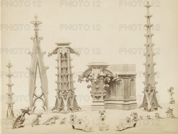 Decorative motifs in copper for Notre-Dame in Rouen, for example gargoyles and leaf motifs, Différents motifs décoratifs en cuivre tortureé pour les Clochetons de la Flèche de la Cathédrale de Rouen, photograph, page, name: 'Witz & cie Phot. Rouen', inscription: 'Exécutés dans les ateliers de F. Marrou, d'après les plans de M. Barthélemy Père', photographer: Witz et Cie., Rouen, c. 1881 - in or before 1884, Witz et Cie., photographer, paper, albumen print, albumendruk, height 215 mm x width 278 mm, piece of sculpture, reproduction of a piece of sculpture, ornament, hybrid animals, Rouen Cathedral, Stone architectural fragments arranged on a flat surface against a plain light background, including tall pointed pinnacles with ornate foliated crockets, a clustered vertical turret with repeating decorative projections, a sculpted corner section with carved arch forms and elaborate leaflike ornamentation, multiple small carved grotesque or birdlike figures positioned on the ground, and vari...