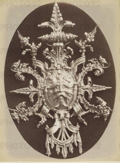 Decorative lead shield with a lion's head and leaf motifs, Cartonche en plomb repoussé an marteau, photograph, page, inscription: 'Composé et exécuté par F. Marrou.', name: 'Witz & cie Phot. Rouen', photographer: Witz et Cie., c. 1880 - in or before 1884, Witz et Cie., photographer, paper, albumen print, height 277 mm x width 208 mm, ornament derived from plant forms, lion's head, ornament, An ornate sculptural relief carved in a pale material is set against a dark oval background, featuring a central stylized human face with a mustache and flowing beard, surrounded by symmetrical arrangements of acanthus leaves, curling ribbons, braided elements, and botanical motifs that radiate outward. The composition includes scroll-like forms, twisted cords, and tassel-like hanging drapery beneath the face, with finely modeled textures and high-contrast lighting that emphasizes the three-dimensional details. The outer edges of the dark oval sit within a lighter rectangular border, and the overall...