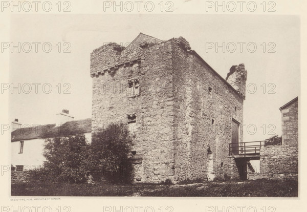 Wraysholme Tower, View of Wraysholme Tower, page, photomechanical print, photographer: Wright, HM & Co., Cumbria, London, c. 1870 - in or before 1880, paper, heliotype, height 111 mm x width 175 mm, tower, clock tower, monumental adornment, A sepia-toned photograph of a stone building with rough textured walls and uneven masonry, showing a tall rectangular tower-like section with small arched window openings and a crenellated top; a pitched roof and a ruined chimney or jagged stone projection rise behind it, while a wooden balcony or walkway with railings connects to the side where a doorway is visible; in front of and beside the building there are bushes and a low stone wall, and adjacent to the main structure a smaller whitewashed house with a sloped roof and chimneys is partially visible.