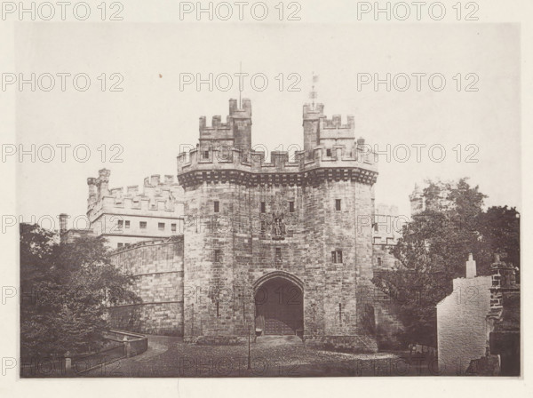 Lancaster Castle. 1876, View of Lancaster Castle, page, photomechanical print, photographer: Wright, HM & Co., Lancaster (Lancashire), London, 1876, paper, photoaquatint, height 127 mm x width 180 mm, castle, gate, entrance, Lancashire, Lancaster Castle, A stone gatehouse with battlements and two projecting round towers frames a large arched entrance, with masonry showing visible blocks and small slit windows; decorative corbels run beneath the parapet and a carved emblem is set above the arch; surrounding buildings with crenellated walls and rectangular windows rise behind the gatehouse; mature trees flank the structure and a curved stone wall and railing lead toward the entrance; a cobbled forecourt and a solitary lamp post stand before the arch, and a partial ruined wall appears at the right edge.
