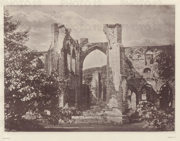 Furness Abbey_the east window, View of the east window of the ruins of Furness Abbey, page, photomechanical print, photographer: Alfred Pettitt, Furness Abbey, Keswick, c. 1870 - in or before 1880, paper, heliotype, height 125 mm x width 170 mm, abbey, monastery, convent, Roman Catholic church, church-window, eg rose-window, Cumbria, A sepia-toned photograph showing the stone remains of a large ruined building with tall arched openings and broken columns, parts of the walls covered in climbing vegetation, leafy trees and bushes framing the left and right sides, a cloudy sky above, and a low stone platform in the foreground.