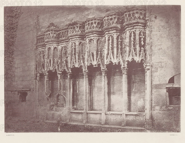 Furness Abbey_the sedilia, Sedilia in the ruins of Furness Abbey, page, photomechanical print, photographer: Alfred Pettitt, Furness Abbey, Keswick, c. 1870 - in or before 1880, paper, heliotype, height 125 mm x width 175 mm, abbey, monastery, convent, Roman Catholic church, parts of church interior, Cumbria, A section of an ornate stone wall with a series of tall, narrow arched niches topped by decorative carved tracery and pinnacles, supported by slender columns and detailed capitals, with weathered surfaces and patches of discoloration; a rough stone floor and a strip of grass run along the base; portions of adjacent plain stone walls frame the carved section on both sides.