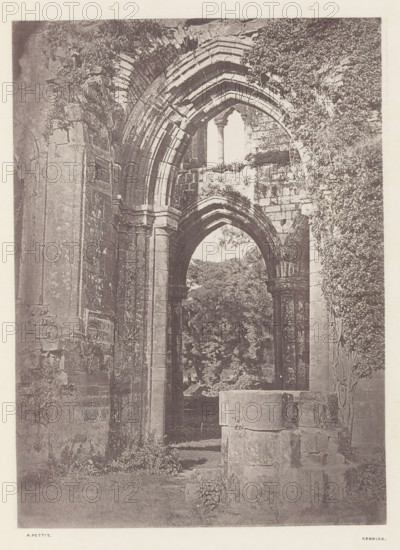 Arch at Furness Abbey, Arch of the ruins of Furness Abbey, page, photomechanical print, photographer: Alfred Pettitt, Furness Abbey, Keswick, c. 1870 - in or before 1880, paper, heliotype, height 169 mm x width 123 mm, arch, archivolt, architecture, ruin of church, monastery, Cumbria, A tall stone archway with layered carved voussoirs frames an opening to a leafy scene beyond, flanked by massive square columns and textured masonry walls showing weathering and patches of vegetation; remnants of another arch and column are visible in front, and climbing plants cover parts of the right wall while rough stone blocks and low grassy ground occupy the foreground.