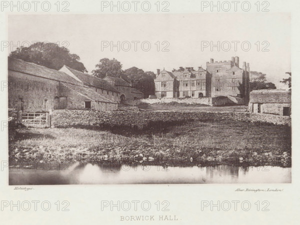 Borwick Hall, View of Borwick Hall, page, photomechanical print, photographer: Alex Rivington, Borwick, London, c. 1870 - in or before 1880, paper, heliotype, height 106 mm x width 175 mm, rural housing, eg country-house, villa, cottage, Lancashire, A wide sepia-toned view shows a large stone manor house with multiple gabled roofs and tall chimneys set behind a low stone wall, flanked by adjoining stone outbuildings with pitched roofs and small doorways; a grassy foreground with scattered rocks slopes down to a reflective body of water at the bottom edge, and a cluster of trees rises behind and between the buildings.