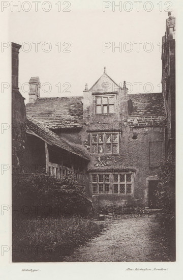 Borwick Hall, View of Borwick Hall, page, photomechanical print, photographer: Alex Rivington, Borwick, London, c. 1870 - in or before 1880, paper, heliotype, height 179 mm x width 117 mm, rural housing, eg country-house, villa, cottage, Lancashire, A tall stone building facade with a pitched roof and multiple leaded windows set in mullioned frames, showing aged and weathered textured walls and roof tiles; a covered wooden gallery or porch projects from the left side with visible timber posts and beams, and dense foliage or shrubs grow along the base of the building beneath the gallery; a gravel or dirt path runs toward the foreground from the building entrance area, and parts of adjacent stone walls and chimneys are visible at the image edges.