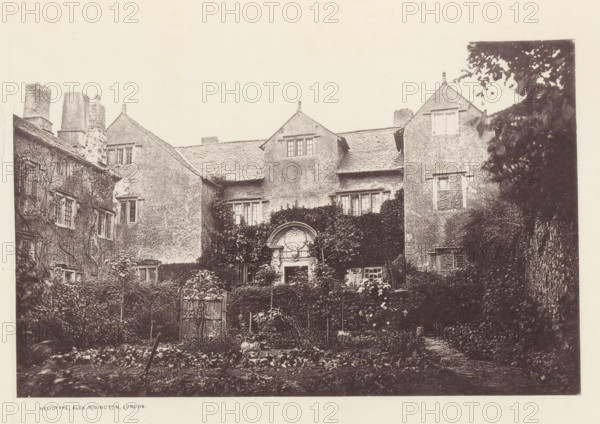 Cark Hall, View of a country house in Cark, page, photomechanical print, photographer: Alex Rivington, Cark, London, c. 1870 - c. 1880, paper, heliotype, height 114 mm x width 180 mm, rural housing, eg country-house, villa, cottage, Cumbria, A sepia-toned photograph of an old multi-story building with textured exterior walls and steeply pitched roofs, featuring dormer windows and tall chimneys; ivy and climbing plants cover portions of the facade and surround a central ornate arched doorway with decorative stonework, while rectangular windows of varying sizes punctuate the walls; in front of the building a garden area with rows of low plants, trellises, and a small wooden gate is visible, and dense foliage frames the right side of the scene.