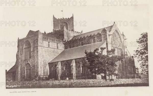 View of the Cartmel Priory, Cartmel church, page, photomechanical print, photographer: Alex Rivington, Cartmel, London, c. 1870 - in or before 1880, paper, heliotype, height 104 mm x width 180 mm, church (exterior), Lancashire, Cartmel Priory, A large stone building with tall arched windows and a peaked roof dominates the image, showing detailed masonry, buttresses, and crenellated parapets; a square tower with a louvered opening and a flagpole rises above the central roofline; trees partially obscure the lower right portion of the structure; a low stone wall runs along the foreground.