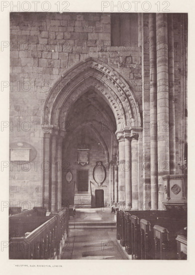Piper choir. Cartmel church, Interior of the Cartmel Priory, page, photomechanical print, photographer: Alex Rivington, Cartmel Priory, London, c. 1870 - in or before 1880, paper, heliotype, height 177 mm x width 128 mm, interior of church, parts of church interior: choir, Lancashire, Cartmel, A stone interior of a building showing a long aisle with wooden pews on either side leading toward an arched doorway; the walls and arch are constructed from large rectangular blocks and the arch features layered, decorative mouldings. Tall cylindrical columns rise along the right side supporting the structure, and a carved stone pulpit with decorative panels is visible near the foreground on the right. Within the archway there are stone steps, recessed niches, and framed oval and rectangular plaques mounted on the wall, with a small door set beneath one of the plaques. The floor shows worn stone paving and shadow patterns cast by the architecture.