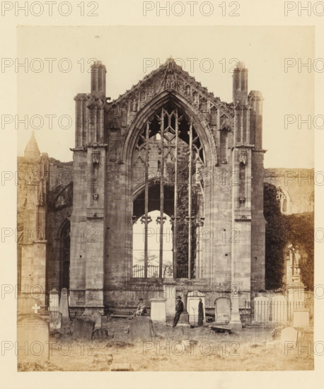 View of a window of the ruins of Melrose Abbey with two men in front of it, photograph, page, title: 'East window', number: '(No. 11.)', inscription: 'Thirty-seven feet in height by sixteen in breadth, at the point of the arch are figures of David I., the Founder of the Abbey, and Matilda, his Queen.', photographer: WJ Pringle, Melrose Abbey, c. 1860 - in or before 1870, Melrose (Scotland), paper, albumen print, height 164 mm x width 141 mm, ruin of church, monastery, church-window, eg rose-window, A sepia-toned photograph of a large Gothic stone façade with a tall pointed arched window featuring ornate tracery and vertical stone mullions, flanked by two tall buttresses and decorative pinnacles; the lower portion shows a graveyard with various upright and leaning headstones, a fenced area, and two men among the monuments, one seated on a bench and the other standing and leaning against a gravestone; ivy or climbing vegetation is visible along part of the right side of the façade and sm...
