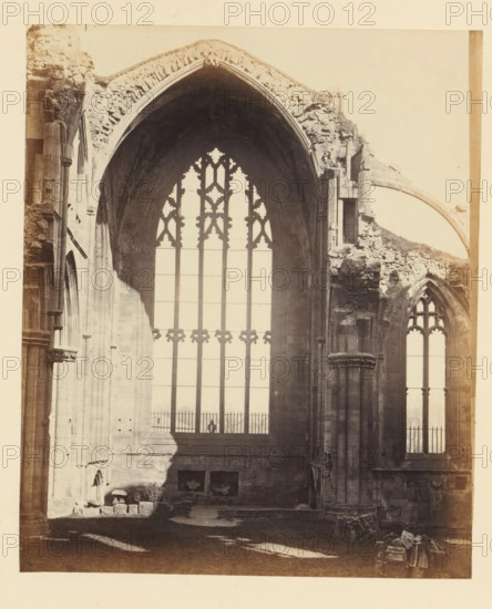 View of a window of the ruins of Melrose Abbey, photograph, page, number: '(No. 12.)', title: 'Interior', inscription: 'Exhibiting the Chancel, and situation of High Altar, with Tomb of Alexander II. of Scotland, and that in wich the Heart of Robert Bruce was deposited.', photographer: WJ Pringle, Melrose Abbey, c. 1860 - in or before 1870, Melrose (Scotland), paper, albumen print, height 165 mm x width 137 mm, ruin of church, monastery, church-window, eg rose-window, A sepia-toned photograph showing the interior of a large stone building with tall arched window openings and ornate tracery, with one central high window framed by a pointed arch and flanked by columns and ruined masonry; portions of the stone walls and archways show weathered and broken edges, rubble and scattered stones lie on the floor near the base of the walls, a shadow falls across part of the wall and floor, and a smaller arched window is visible to the right with Gothic tracery.