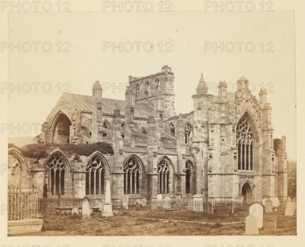 View of the ruins of Melrose Abbey, photograph, page, number: '(No. 1.)', title: 'The Abbey from the south-west', photographer: WJ Pringle, Melrose Abbey, c. 1860 - in or before 1870, Melrose (Scotland), paper, albumen print, height 134 mm x width 173 mm, ruin of church, monastery, A sepia-toned photograph of a large stone Gothic-style building with arched windows and ornate tracery, sections of the façade showing pointed arches and tall narrow windows, buttresses and decorative pinnacles, and a partially ruined archway on one side; in front of the building there is a grassy area with fenced enclosures, stone tombstones and monuments including an obelisk, and wrought iron railings surrounding some graves.