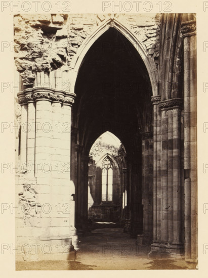 View of an aisle of the ruins of Melrose Abbey, photograph, page, title: 'Fine perspective view of south aisle', number: '(No. 4.)', inscription: 'The Carving and Capitals of Pillars, remarkable', photographer: WJ Pringle, Melrose Abbey, c. 1860 - in or before 1870, Melrose (Scotland), paper, albumen print, height 171 mm x width 121 mm, ruin of church, monastery, parts of church interior: aisle, A tall stone archway framed by thick columns and ornate capitals leads into a dark vaulted interior with layered arches receding toward a distant pointed window, rough stone walls and carved column shafts visible on either side, light filtering through the far window creating contrasts of shadow and pale stone on the floor and lower walls.