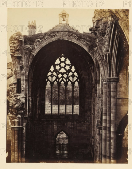 Interior with a view of a window of the ruins of Melrose Abbey, photograph, page, number: '(No. 5.)', inscription: 'With Belfry and Font to the right.', title: 'Interior of south transept', photographer: WJ Pringle, Melrose Abbey, c. 1860 - in or before 1870, Melrose (Scotland), paper, albumen print, height 175 mm x width 138 mm, ruin of church, monastery, church-window, eg rose-window, A tall stone arch with a tracery window sits between massive ribbed columns and weathered masonry, the window opening filled with ornate quatrefoil and trefoil shapes revealing trees and a fence beyond, rough stonework and carved capitals flank the arch on both sides, an arched doorway with a gate is visible below, broken and crumbling sections of the structure show exposed layers of stone and patches of vegetation, and a small bell tower rises above the archline against a pale sky.