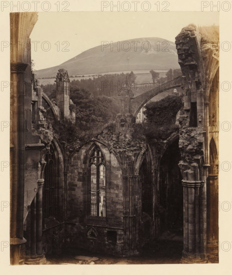 View of a chapel of the ruins of Melrose Abbey with Eildon Hill in the background, photograph, page, title: 'Chapel to east of south transept', number: '(No. 6.)', inscription: 'With Tomb of Michael Scott, the Wizard, and Eildon Hill in the distance.', photographer: WJ Pringle, Melrose Abbey, c. 1860 - in or before 1870, Melrose (Scotland), paper, albumen print, height 169 mm x width 140 mm, ruin of church, monastery, (high) hill, parts of church interior: chapel, A sepia-toned photograph shows the ruins of a stone building with tall, arched windows and broken, weathered columns and lintels, revealing jagged edges and crumbling masonry; vegetation grows among the ruins and over some upper sections, with leafy trees visible beyond the structure; in the background a gently sloping hill rises behind a patchwork of fields and lines of trees, and an arched stone bridge or buttress spans across part of the scene.