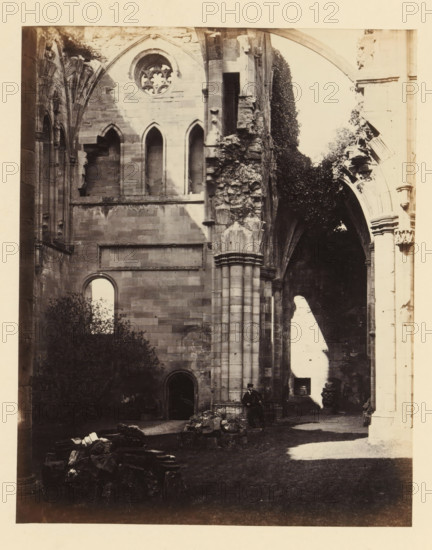 Interior of the north transept of the ruins of Melrose Abbey, photograph, page, title: 'Interior view of north transept', inscription: 'On the left, Statue of St. Peter with the Keys, in front, Crown of Thorns Window.', number: '(No. 7.)', photographer: WJ Pringle, Melrose Abbey, c. 1860 - in or before 1870, Melrose (Scotland), paper, albumen print, height 165 mm x width 131 mm, ruin of church, monastery, parts of church interior: transept, A sepia-toned photograph of a ruined stone building interior showing tall arched openings and a round tracery window high on a wall, with sections of carved columns and crumbling masonry. Ivy or dense foliage grows over part of the ruins near one of the arches. Sunlight creates a bright patch on the floor and a contrasting shadow on the wall. Rubble and scattered stone fragments lie on the ground in the foreground. A man wearing dark clothing and a hat is seated at the base of a large column near the center right of the image.