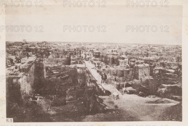View of a diamond mine in South Africa, presumably near Kimberley, page, photomechanical print, number: 'Pl. 1', photographer: Gray Brothers (possibly), South Africa, c. 1870 - in or before 1880, paper, collotype, photogravure, height 123 mm x width 185 mm, Print in rear image section., mine, Kimberley, A wide sepia-toned photograph showing a rough landscape of deep, irregular trenches and mounds of earth stretching toward the horizon, with narrow plank walkways bridging the gaps between elevated dirt walls and scattered wooden supports and ladders set among the excavated areas; small groups of figures in dark clothing stand or walk along the paths and between the trenches, and various indistinct tools and debris lie on the ground; the sky appears pale and empty above the scene, while the foreground features steep vertical edges of the trenches and rough, textured soil.