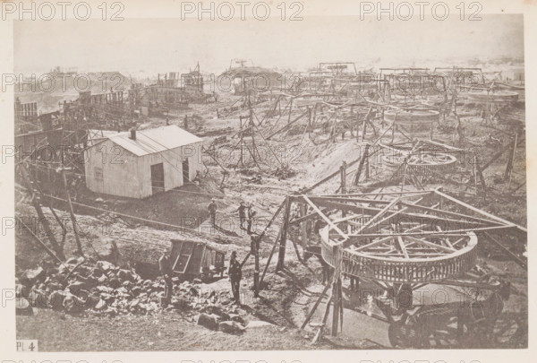 View of a diamond mine in South Africa, presumably near Kimberley, page, photomechanical print, number: 'Pl. 4', photographer: Gray Brothers (possibly), South Africa, c. 1870 - in or before 1880, paper, collotype, photogravure, height 125 mm x width 192 mm, Print in rear image section., mine, Kimberley, A wide sepia-toned photograph shows an industrial site with rows of circular wooden structures on raised frames and triangular timber supports scattered across a rough, dirt-covered landscape. In the foreground a cluster of round wheel-like frames sits atop latticed bases, with angled beams and poles extending outward. A small corrugated building with a sloped roof and an open doorway stands to the left of center, connected to the rest of the site by thin wires or ropes. Several men in dark clothing and hats stand and walk along muddy paths between the structures, and various wooden platforms, scaffolding, and piles of rocks or rubble are visible across the scene. The background fades i...
