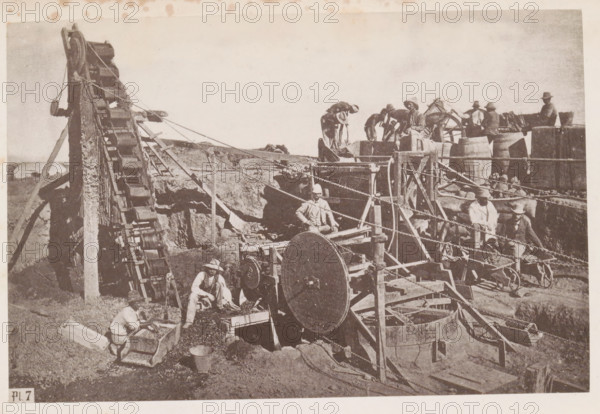 Men at work in a diamond mine in South Africa, presumably near Kimberley, page, photomechanical print, number: 'Pl. 7', photographer: Gray Brothers (possibly), South Africa, c. 1870 - in or before 1880, paper, collotype, photogravure, height 125 mm x width 189 mm, Print in rear image section., mine, Kimberley, A sepia-toned scene shows a group of people working among wooden machinery and barrels in an open, dusty area. A large inclined wooden conveyor with rectangular buckets runs up the left side, supported by a tall frame and ropes. In the center foreground a large round wooden wheel is mounted within a wooden structure with beams and pulleys, and ropes and chains run across the scene connecting various components. Several men wearing wide-brimmed hats and light clothing are positioned around the machinery; some stand on raised platforms near barrels while others crouch or sit at workstations handling material in wooden troughs and carts. Stacked barrels and wooden crates appear near...