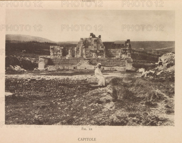 Capitole, View of a Roman ruin at Lambaesis in Algeria, page, photomechanical print, number: 'Fig. XX', Algeria, maker: Berthaud Frères, France, c. 1884 - in or before 1894, photographer, North Africa, paper, collotype, photoprint, height 108 mm x width 158 mm, ruin of a building, architecture, Lambaesis, A sepia-toned photograph of an ancient stone ruin set in a grassy, uneven landscape, showing large rectangular stone blocks forming partial walls and a raised platform with a few standing columns and column fragments; a figure draped in a light-colored robe is seated on a stone ledge in the foreground facing the ruins; scattered stones and rubble lie around the structure, and the background reveals low rolling hills and sparse vegetation under a pale sky.