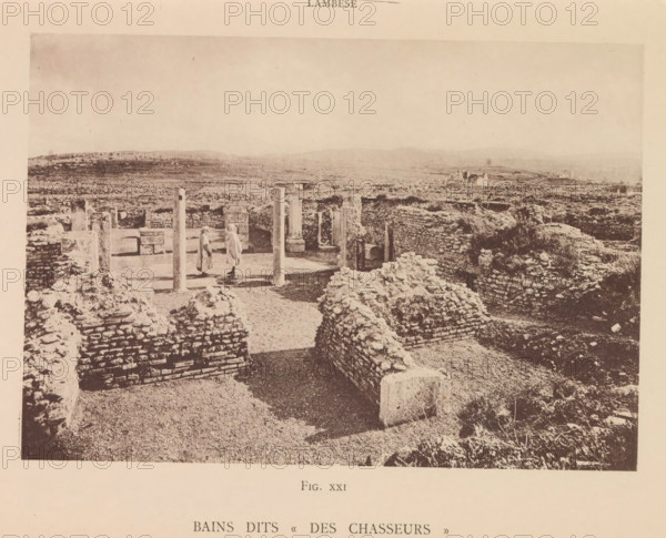 View of the ruins of a Roman bathhouse at Lambaesis in Algeria, Bains dits Des Chasseurs, page, photomechanical print, number: 'Fig. XXI', Algeria, maker: Berthaud Frères, France, c. 1884 - in or before 1894, photographer, North Africa, paper, collotype, photoprint, height 102 mm x width 147 mm, ruin of a building, architecture, public baths, Lambaesis, A wide, sepia-toned photograph shows an archaeological site with low stone walls and rubble forming rectangular enclosures across a flat, open landscape. In the foreground are rough stone mounds and short brick-like walls leading toward an inner courtyard area framed by upright columns and partial walls. Two figures wearing long garments stand near the columns within the courtyard. Beyond the ruins the land stretches to gently rolling hills under a pale sky, and scattered isolated structures appear in the distance. The image has a border of blank paper around it.