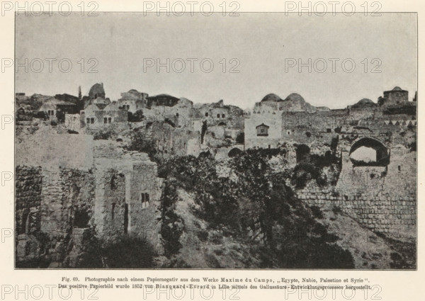 Adjutant des Fürsten Danilo von Montenegro. Nach einem Papiernegativ von P. Jovanovits, Belgrad (1850 or 1852), View of Jerusalem, page, photomechanical print, number: 'Fig. 69.', inscription: 'Photographie nach, hergestellt.', photographer: Maxime Du Camp, Jerusalem, cliché maker: anonymous, c. 1900 - in or before 1905, paper, autotypie, height 115 mm x width 118 mm, city-view in general, 'veduta', A wide monochrome photograph of an old stone settlement showing numerous ruined and intact masonry structures spread across a rocky hillside under a pale sky; visible are blocky buildings with flat and domed roofs, arched openings, small rectangular windows, and sections of crumbling walls and terraces; vegetation grows among the stones and along the slopes, and a prominent archway stands near the foreground beside stacked stone courses.