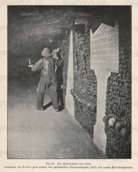 Die Catacomben von Paris, Unknown man in the Catacombs of Paris, page, photomechanical print, number: 'Fig. 98.', photographer: Félix Nadar, Catacombs of Paris, cliché maker: anonymous, c. 1900 - in or before 1905, Paris, paper, autotypie, height 134 mm x width 108 mm, adult man, catacombs, A grainy black-and-white scene showing a dim underground passage with walls lined by stacked human skulls and bones, a person wearing loose clothing and a wide-brimmed hat facing the wall and holding a candle or small torch in one hand while appearing to inspect the bones, and a tall marker or plaque positioned among the ossified walls.