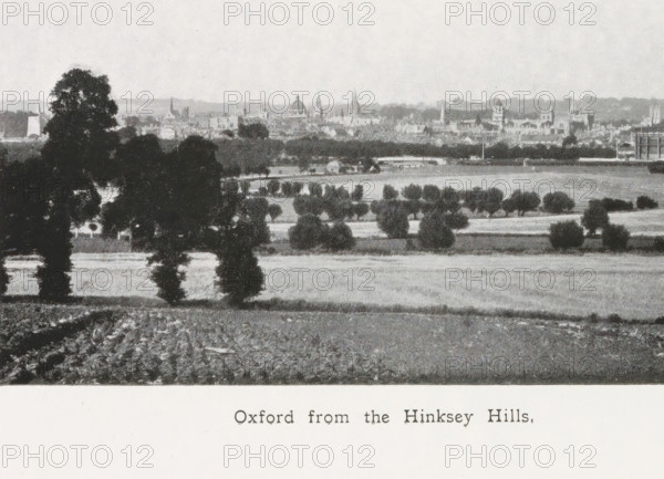 Oxford from the Hinksey Hills, page, photomechanical print, inscription: ''That sweet city with her dreaming spires.'', maker: Henry W. Taunt, Oxfordshire, c. 1907 - in or before 1912, paper, autotype, height 79 mm x width 132 mm, winter landscape, landscape symbolizing winter (the four seasons of the year), Oxford, A vertical black and white photograph of a landscape is visible on the left side, showing trees, fields, and distant buildings with a skyline; the photograph has a narrow white border and is rotated ninety degrees. The rest of the image shows a book page with closely spaced printed lines of text occupying the right side and lower portion, and a visible center gutter where the pages meet. The overall tones are grayscale with paper textures and slight shadows along the page crease.