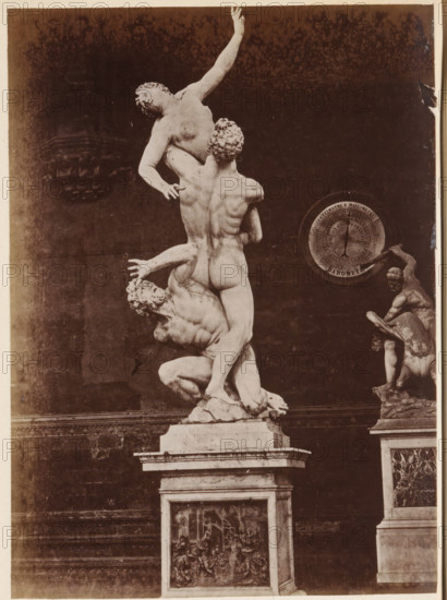 The Rape of the Sabine Virgins by Giambologna, photograph, page, Italy, c. 1850 - in or before 1860, photographer, paper, albumen print, height 132 mm x width 96 mm, piece of sculpture, reproduction of a piece of sculpture, rape of the Sabine women: they are carried off by Romulus and his men during a feast, A photograph of a large marble sculpture showing three intertwined nude figures on a rectangular pedestal, with one figure lifted high and reaching upward while another supports from below and the third crouches at the base, detailed muscular forms and curly hair visible; to the right, a smaller statue of two figures on a separate pedestal is partially visible, and a circular dial-like object is mounted on the wall behind them; the background is a dark wall with some lighter mottling and the pedestals show carved relief panels.