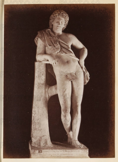 Resting Satyr by Praxiteles in the Capitoline Museums, photograph, page, Rome, c. 1850 - in or before 1860, photographer, paper, albumen print, height 134 mm x width 98 mm, piece of sculpture, reproduction of a piece of sculpture, Capitoline Museums, A photograph of a marble statue of a nude male figure leaning against a tree-like support, the figure shown from head to feet with curly hair and a relaxed facial expression, a draped cloth across one shoulder and over part of the torso, one arm resting on the support and the other hand placed near the hip, a small leaf placed over the groin, the statue standing on a rectangular base against a dark background.