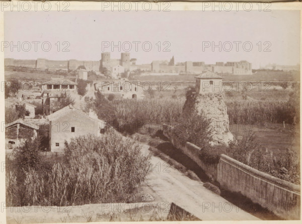 View of the Via Appia with the Porta San Sebastiano in Rome in the distance, photograph, page, Rome, c. 1850 - in or before 1860, photographer, paper, albumen print, height 97 mm x width 134 mm, Photo after page 222., street, landscape with ruins, city-gate, Via Appia Antica, A sepia-toned landscape showing a dirt road leading toward a cluster of small buildings with tiled roofs surrounded by tall grasses and vegetation, a low wall running beside the road, a square tower-like structure on a raised, rough stone base near the right side, additional houses and farm structures scattered among fields and reeds, and a distant fortified wall with towers and crenellations along the horizon under a pale sky.