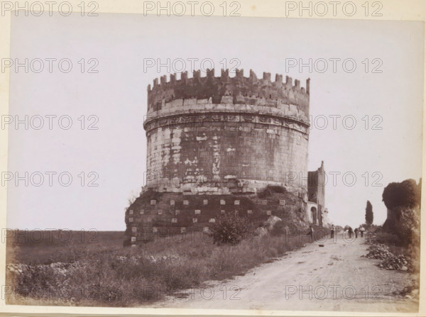 View of the Tomb of Caecilia Metella on the Via Appia near Rome, photograph, page, Via Appia Antica, c. 1850 - in or before 1860, photographer, paper, albumen print, height 97 mm x width 135 mm, Photo after page 222., street, landscape with ruins, grave, tomb, A large circular stone tower with a crenellated top stands on a grassy mound beside a dirt road, the tower showing weathered stone blocks and a decorative band near its upper section; small figures walk and stand along the road near the tower, and a tall narrow tree and a larger tree appear further along the path, with low vegetation and scattered rocks in the foreground.