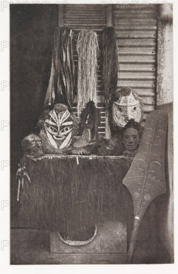 Masks and a paddle in Tahiti, Various masks, paddle, &c., page, photomechanical print, number: 'No. 23.', photographer: Henry Stuart-Wortley, Tahiti, cliché maker: Meisenbach's Autotype Company, printer (person): Meisenbach's Autotype Company, 1880, Meisenbach's Autotype Company, photographer, French Polynesia, paper, collotype, photoprint, height 168 mm x width 105 mm, mask, A vertical image showing a display of carved masks and woven materials against a textured background. At the top center several bundles of long strands hang down, varying in thickness and texture; a shutter and corrugated surface are visible behind them. Below the hanging strands are multiple carved faces and masks with distinct shapes and painted or carved facial patterns, some with hairlike material attached around them. A flat surface covered in a rough, grasslike fringe supports the masks and other small carved objects, and a carved elongated shield with patterned incisions leans against the right side. The le...