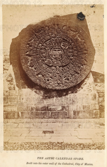 The Aztec calendar stone, photograph, page, inscription: 'Built into, City of Mexico.', Mexico City, c. 1870 - in or before 1880, photographer, North America, paper, albumen print, height 142 mm x width 100 mm, Photo on frontispiece., church (exterior), calendar, almanac, Aztecs, A large circular carved stone disk with intricate radial patterns and raised relief motifs is mounted against a rough stone wall; the disk shows concentric rings filled with geometric and figurative carvings and a central face-like figure, with serpent-like designs near the lower edge. The surrounding wall is composed of rectangular stone blocks with visible mortar lines and weathering, and a paved stone ground extends outward below the wall. The photograph has a sepia tone and shows some surface blemishes and discoloration on the wall and disk.