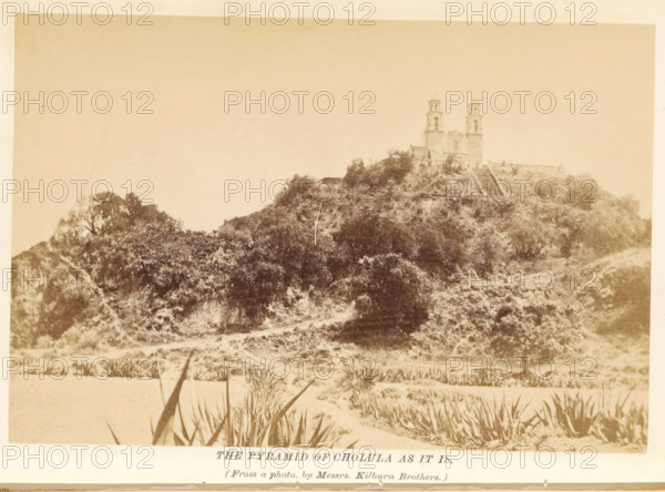 The pyramid of Cholula as it is, photograph, page, photographer: Kilburn, Mexico, c. 1870 - in or before 1880, North America, paper, albumen print, height 102 mm x width 151 mm, Photo after page 156., church (exterior), quadrangular pyramid, Great Pyramid of Cholula, Iglesia de Nuestra Señora de los Remedios, A sepia-toned photograph showing a large earthen mound covered in vegetation with a path or cleared area at its base and spiky plants in the foreground, and a stone building with twin towers situated atop the mound, stair-like terraces or steps leading up the slope, scattered trees and shrubs on the hillside, and a plain sky above.