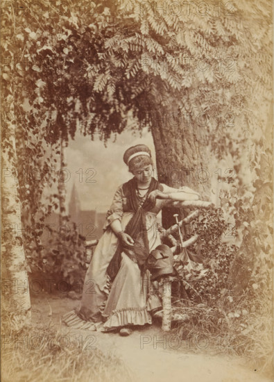 You Ring on my Finger, My gold ring, Portrait of an unknown woman sitting on a bench in front of a tree, presumably looking at a ring on her finger, Frauen-Liebe und Leben, photograph, page, number: '4.', name: 'Photog. und Verlag v. H. Hirsch, Berlin, N Christinenstr. 36 & 37', photographer: Hugo Hirsch, 1880, paper, albumen print, albumendruk, height 134 mm x width 95 mm, adult woman, bench, ring, A sepia-toned scene of a young woman leaning against a rough wooden fence beneath overhanging leafy branches, wearing a long dress with ruffled hem and layered shawls, her hair wrapped with a headband and her gaze directed downward, one arm resting on the fence and the other hand holding fabric, a hat and a walking stick hanging from the fence beside her, tree trunks and dense foliage surrounding the path behind her.