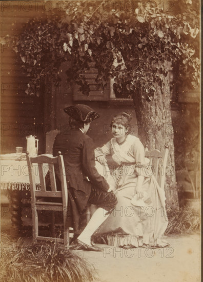 Süsser Freund, du blickest mich verwundert an, Kannst es nicht begreifen, wie ich weinen kann, Portrait of an unknown woman and man, sitting at a table in a garden, Frauen-Liebe und Leben, photograph, page, name: 'Photog. und Verlag v. H. Hirsch, Berlin, N Christinenstr. 36 & 37', number: '6.', photographer: Hugo Hirsch, 1880, paper, albumen print, albumendruk, height 134 mm x width 95 mm, garden, adult woman, adult man, historical costume, A sepia-toned scene of two people seated outdoors beneath a tree with dense leafy branches, one person facing away wearing a dark coat and a tricorne-style hat, the other facing forward wearing a light-colored dress with ruffled sleeves and a sash, the forward-facing person resting an arm on the other's knee and holding a folded cloth in the other hand, wooden chairs with simple carved backs and a small table covered with a fringed cloth visible beside them, a pitcher and a glass on the table, a tree trunk rising behind them, grass clumps at the for...