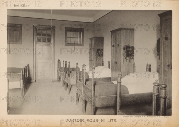 Dortoir pour 16 lits, Dormitory in a barracks of the Svea livgarde in Stockholm, page, photomechanical print, number: 'N:o XII.', maker: V. Wolfenstein, Stockholm, maker: Johannes Jaeger, c. 1888 - in or before 1891, paper, collotype, photoprint, height 120 mm x width 187 mm, bed, A sepia-toned photograph of a dormitory interior showing a row of wooden beds with turned posts and plain headboards aligned along the right side, each bed made with a blanket and a pillow. Along the far wall are tall wooden wardrobes with visible grain and hardware, some with leather bags or straps hanging from their doors. A closed paneled door and windows with muntins are set into the left and back walls, and two vertical cords hang from the ceiling near the windows. The floor is bare and extends toward the foreground, and a narrow strip of wall and ceiling molding runs around the room. The image is bordered by a lighter margin.