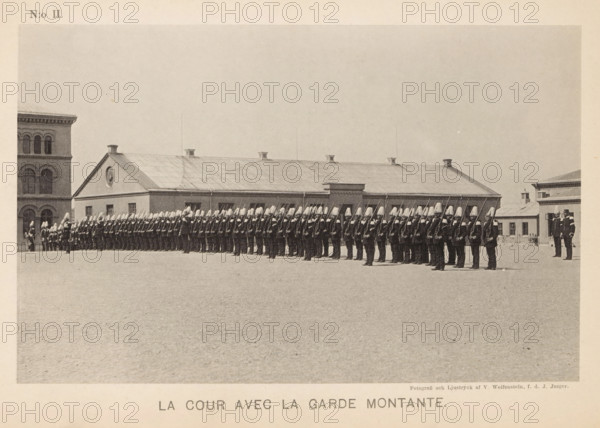 Courtyard with members of the Svea livgarde, La cour avec la garde montante, page, photomechanical print, number: 'N:o II.', maker: V. Wolfenstein, Stockholm, maker: Johannes Jaeger, c. 1888 - in or before 1891, paper, collotype, photoprint, height 118 mm x width 186 mm, courtyard, guard of honor, A wide courtyard scene shows a long line of uniformed guards standing in formation in front of a low building with a pitched roof and small chimneys, the guards wearing dark uniforms and tall light-colored helmets and holding long rifles upright, with a taller multi-story building visible at the left edge and another building at the right edge, the ground in the foreground appearing flat and textured and the sky above pale and empty.