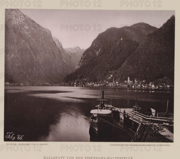 View of Hallstatt, Hallstatt von der Eisenbahn-haltestelle, page, photomechanical print, number: 'III.', photographer: Friedrich Simony, Salzburg (city), cliché maker: Friedrich Bruckmann, publisher: Eduard Hölzel, Vienna, 1886, Lower Austria, paper, collotype, photoprint, height 147 mm x width 187 mm, mountain-range, snow peaks, Upper Austria, Hallstatt, A sepia-toned scene showing a calm body of water bordered by steep, forested mountains and rocky cliffs, with a small lakeside village nestled at the base of the hills, visible church steeples and clustered buildings along the shoreline, a wooden dock in the foreground with moored boats and a pier extending into the water, and reflections of the shoreline and mountains faintly visible on the water surface.