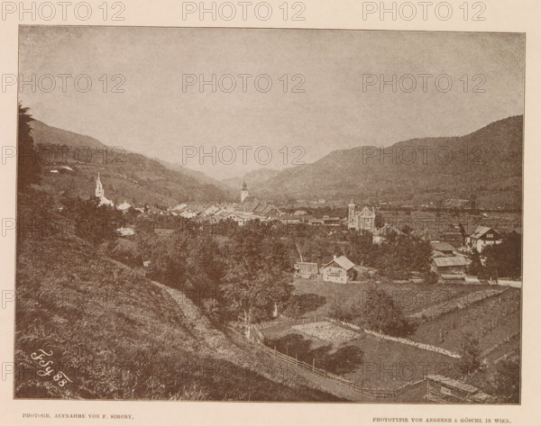 View of Schladming, Östliche Ansicht von Schlädming, page, photomechanical print, number: 'IV.', photographer: Friedrich Simony, Salzburg (city), cliché maker: Angerer & Göschl, C., Vienna, publisher: Eduard Hölzel, 1888, Lower Austria, paper, autotypie, height 145 mm x width 195 mm, mountain-range, snow peaks, Steiermark, Schladming, A sepia-toned landscape photograph showing a valley with rolling hills and a village nestled between them, small clustered buildings and houses with pitched roofs spreading across the middle ground, a tall church tower and another steepled structure rising above the rooftops, open fields and fenced plots on the nearer slopes, a winding dirt path leading down the hillside, scattered trees and patches of vegetation across the scene, distant layered hills forming a backdrop under a broad, pale sky.