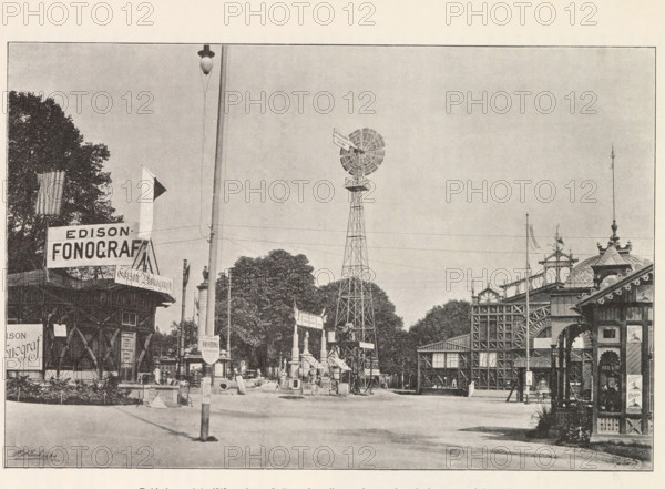 View of the Výstavište in Prague with a building for phonographs, Pohled after výstaviste your stromoradi mezi pavilonem fonografu a budovou pro inzenýrstvi, page, photomechanical print, Výstavište, cliché maker: Carl Bellmann, Prague, c. 1888 - in or before 1893, photographer, paper, autotypie, height 175 mm x width 262 mm, temporary exhibition, 'Salon', façade (of house or building), A wide street scene shows several ornate pavilions and wooden structures with decorative trim and latticework along the sides, a tall metal windmill tower with a multi-bladed fan near the center, a tall pole with a streetlamp near the foreground, clusters of trees behind the buildings, a flag on a pole and another small triangular pennant visible, various signs mounted on buildings and a banner above an entrance, scattered posts and small market-like objects on the ground, and a mostly clear sky occupying the upper portion of the image.