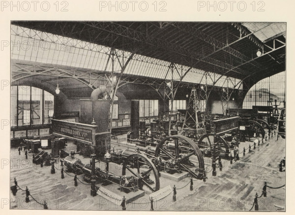 Instalace strojovny. Pohled smear k vychodnimu vchodu, Interior of an engine room, page, photomechanical print, Prague, cliché maker: Carl Bellmann, c. 1890 - in or before 1895, photographer, paper, autotypie, height 176 mm x width 258 mm, machines, parts of machines, tools and appliances, A large indoor industrial space with a high arched roof supported by metal trusses and columns, rows of tall windows along the upper walls, and hanging light fixtures. The floor area contains multiple large mechanical machines with visible flywheels, pistons, and belts, arranged on raised platforms and surrounded by short posts connected with chains. There are several tall structural towers or frames among the machinery and a few small display boards or panels mounted on walls. The overall scene is monochrome with a grainy texture and shows architectural details of the roof, beams, windows, and the dense arrangement of industrial equipment.