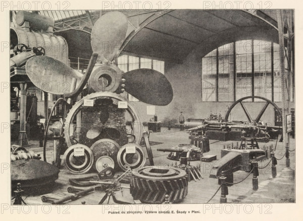 Pohled do strojovny. Výstava závodu F Ringhoffra na Smichove, Interior of an engine room with an exhibition of Skoda, page, photomechanical print, Prague, cliché maker: Carl Bellmann, c. 1890 - in or before 1895, photographer, paper, autotypie, height 176 mm x width 258 mm, interior, representation of a building, machines, parts of machines, tools and appliances, A large industrial interior with tall windows and arched roof supports contains heavy machinery and large metal components arranged across the floor; a prominent multi-bladed propeller is mounted near the center surrounded by circular gears and ring-shaped parts, with various shafts, pistons, and mechanical housings visible nearby; columns and structural beams rise toward the ceiling, and a large flywheel sits toward one side; workbenches, stands, and scattered smaller tools occupy the foreground and background, and the scene is rendered in a monochrome, grainy photographic style.