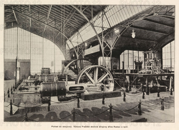 Interior of a machine room. Interior of the Ruston mechanical drive company in Prague, Interior of a machine room with an exhibition of the Ruston mechanical drive company in Prague, page, photomechanical print, Prague, cliché maker: Carl Bellmann, c. 1890 - in or before 1895, photographer, paper, autotype, height 176 mm x width 258 mm, interior, representation of a building, machines, parts of machines, tools and appliances, A large indoor industrial space with a high arched roof and exposed metal trusses, tall windows along the back and side walls, rows of heavy stationary machinery and large flywheels arranged on the floor, metal railings and posts with ropes forming barriers around the equipment, a raised platform supporting additional mechanical apparatus and piping, hanging light fixtures from the ceiling, a few standing figures near the machines, textured floor surfaces and shadow patterns cast by the posts and ropes.