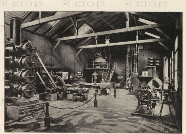Pohled do vnitr pavilonu firmy Novak a Jahn v Praze, Interior of an engine room with an exhibition of Novak and Jahn, page, photomechanical print, Prague, cliché maker: Carl Bellmann, c. 1890 - in or before 1895, photographer, paper, autotypie, height 176 mm x width 258 mm, interior, representation of a building, machines, parts of machines, tools and appliances, A large industrial interior with exposed wooden roof beams and a high ceiling, filled with a variety of heavy machinery and mechanical apparatus. On the left, a vertical stack of cylindrical components sits beside a large flywheel and belt assembly, with angled support beams leaning against them. In the center, a raised platform with a domed vessel and a curved staircase leading up to it is visible, with hanging cables and a suspended light fixture above. Around the floor are multiple engines, pumps, and mechanical linkages mounted on bases and short pedestals, connected by pipes and belts. On the right, tall vertical columns ...