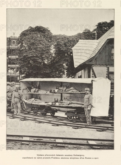 Sleeping compartment on rails, Výstava prenosných zeleznic soustavy Dolbergovy, uspodaradaná ve volné prostore Prazskou akciovou strojirnou drive Ruston a spol, page, photomechanical print, Czech Republic, cliché maker: Carl Bellmann, Prague, c. 1891 - in or before 1895, photographer, paper, autotypie, height 222 mm x width 171 mm, bed, railway-coach, railway-carriage, wagon, A sepia-toned photograph showing a rail setting with tracks in the foreground and a covered wagon-like stretcher platform on small rail carts holding people lying down under a canopy; several standing men in uniform are positioned around the platform, one near the front partially obscuring the end of the canopy, and others grouped toward the left side; behind them a timber-framed building with a steep, shingled roof occupies the right background while large leafy trees fill the central background; to the left a tall wooden tower structure rises above the trees.