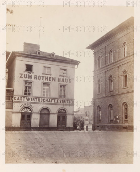 Square in Mainz, photograph, page, photographer: B. Erdmann, Mainz, c. 1867 - in or before 1872, Rhineland-Palatinate, paper, albumen print, height 186 mm x width 152 mm, village square, Place, A street scene showing two adjacent multi-story buildings separated by a narrow passage and framed by a broad empty pavement in the foreground. The left building has a smooth facade with rectangular and arched windows, a pitched roof with small dormers and a chimney, and three arched doorways at ground level with decorative fanlights above two of them. The right building has a masonry facade with regularly spaced arched windows on both visible floors, a prominent cornice with bracketed eaves, and a raised stone base. Between the buildings, a small open area reveals a statue on a pedestal and some ornamental lamp posts, with a few potted shrubs near the entrance to the passage.
