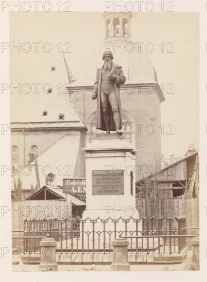 Monument to Johannes Gutenberg in Mainz, photograph, page, number: '15', photographer: B. Erdmann, Mainz, c. 1867 - in or before 1872, Rhineland-Palatinate, paper, albumen print, height 208 mm x width 150 mm, monument, statue, A tall stone pedestal stands behind a decorative metal fence, supporting a life-size bronze statue of a bearded man wearing a long coat and hat, with one hand holding a rolled object against his chest and the other hanging by his side, set against the façade of a large church with a steep roof, arched windows, and a domed bell tower visible above the statue, while wooden buildings and construction materials occupy the area around the base of the pedestal.